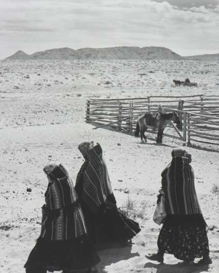 Three people in traditional clothing walk across a sandy landscape toward a wooden corral with a horse inside; hills and cloudy sky in the background.