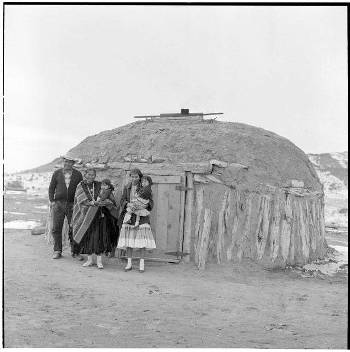 Four people stand in front of a traditional earth and wood structure, known as a hogan, in a sparsely vegetated landscape.