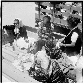 Five women wearing headscarves sit in the back of a truck, sharing a meal with food and drinks placed on the floor.