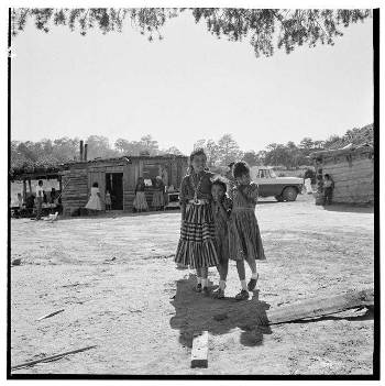 Three girls stand together in front of a dirt yard with wooden shacks and a parked car in the background. Other people and trees are visible further back.