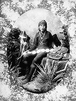 Black-and-white portrait of a seated Native American man wearing traditional clothing and jewelry, with plants and a horse in the background.
