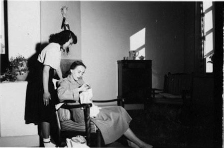 A seated woman is sewing while another woman stands beside her in a sparsely furnished room with sunlight streaming through a window.