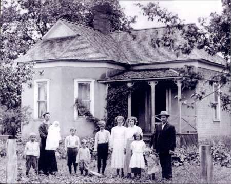 A black-and-white photo of a family with seven children and two adults standing in front of a small house with a porch and sloped roof, surrounded by trees and plants.