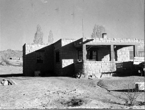 A small, single-story stone house with a flat roof and covered porch sits in a barren, dirt yard under clear skies.