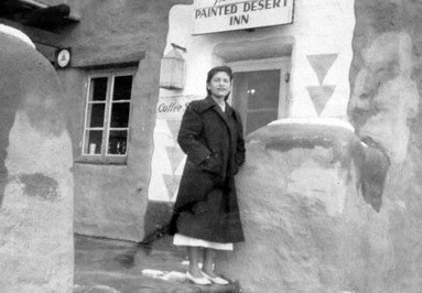A woman in a dark coat stands in front of the Painted Desert Inn, next to stucco walls and a window, in a black-and-white photo.