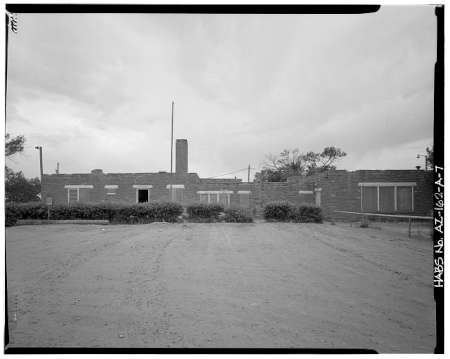 Black and white photo of a single-story brick building with several windows, a central chimney, and a dirt lot in the foreground. Shrubs line the front of the building.