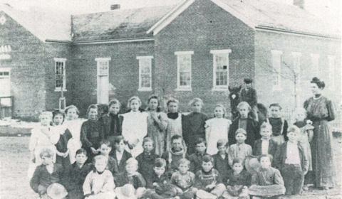 A black-and-white photo of a group of children and two adults posed in front of a brick school building.