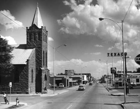 Black and white photo of a small town street with a stone church on the left, cars parked along the road, and Texaco gas station signs visible on the right.