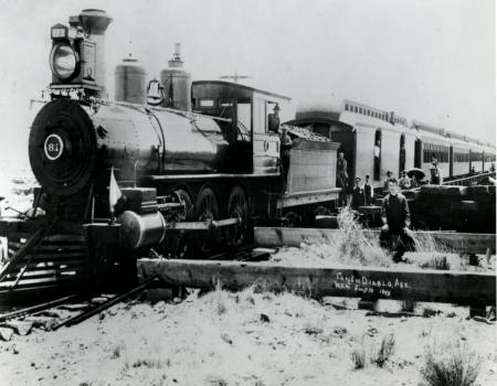 Black and white photo of a steam locomotive stopped near damaged tracks, with a man standing beside debris and train cars in the background.