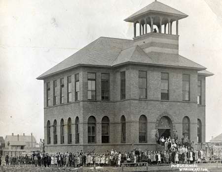 Black-and-white photo of a two-story brick school building with a bell tower, with a large group of people gathered outside the entrance.