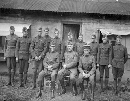 A group of thirteen soldiers in uniform pose for a photo outside a rustic building; three are seated in front and the rest stand behind them.
