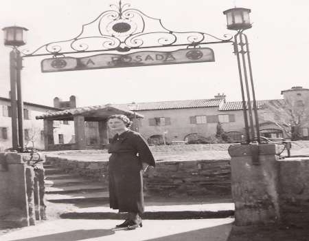 A woman stands under a wrought iron sign reading "LA POSADA" at the entrance of a courtyard with old-style buildings in the background.