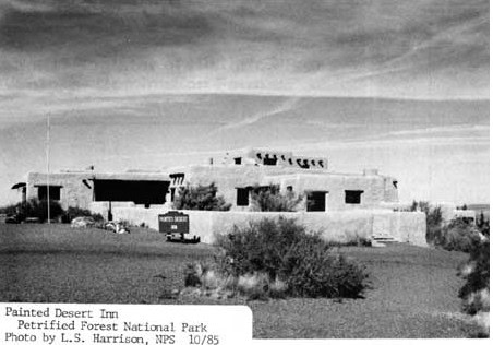 Black and white photo of the Painted Desert Inn, an adobe building in Petrified Forest National Park, surrounded by desert vegetation under a sky with streaked clouds.
