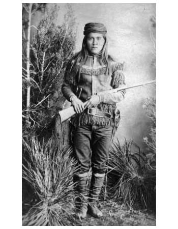 A person in fringed frontier clothing and boots stands holding a rifle, surrounded by foliage in a black-and-white studio portrait.