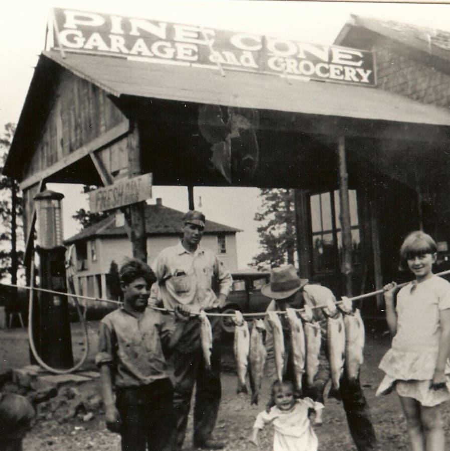 Four children and one adult pose in front of Pine Cone Garage and Grocery, holding a line of large fish; an old gas pump is visible on the left.