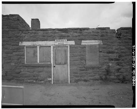 Black-and-white image of a stone building with boarded windows and a sign above the door reading “Principal’s Office.” The building appears weathered and abandoned.
