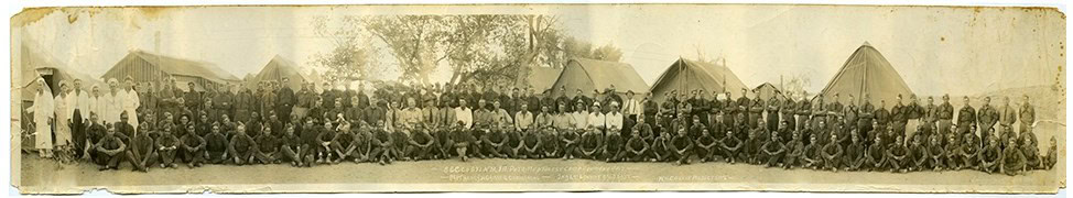 A large group of men, some seated and some standing, pose in front of tents and trees in an outdoor setting, possibly a historical military camp.