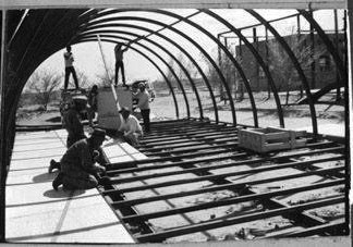 Several people work on constructing a metal-framed structure, assembling the framework and floor panels outdoors under daylight.