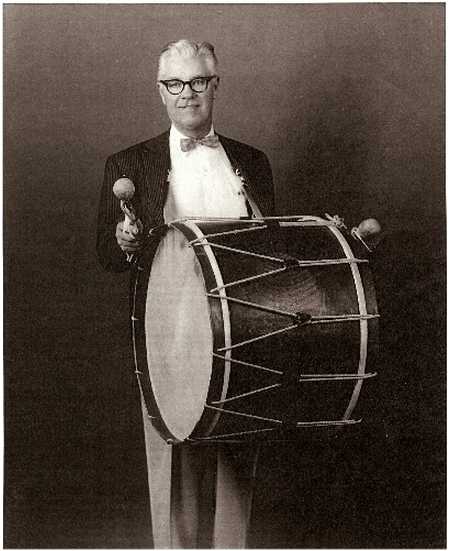 A man wearing glasses and formal attire stands holding a large bass drum and two mallets against a plain background.