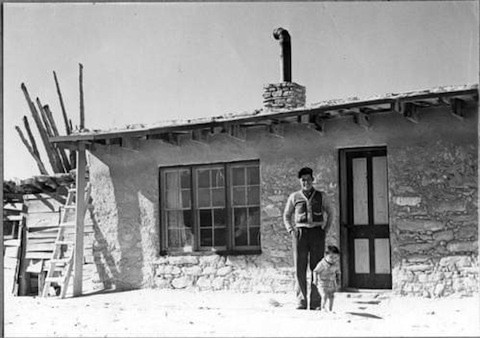 A man and child stand in front of a small, rustic stone house with a chimney pipe, wooden windows, and a simple porch in a rural setting.