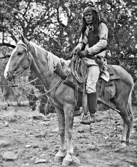 A man with long hair wearing traditional clothing sits on a saddled horse outdoors with trees in the background.