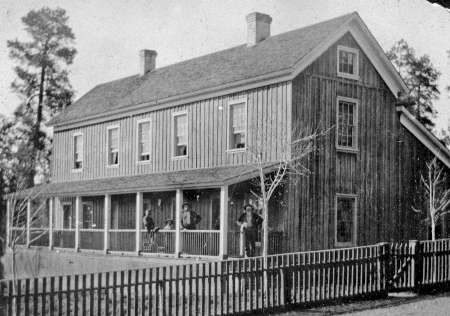 A black and white photo of a two-story wooden building with a porch, fenced yard, and several people standing on the porch.