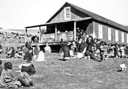 A group of Native American people gather outside a wooden building on open land, some sitting or standing in blankets, with more people dispersed in the background.