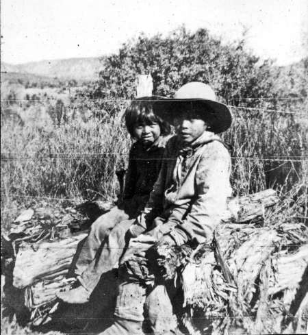 Two children sit on a wooden fence outdoors, with brush and hills visible in the background. One child wears a wide-brimmed hat. The image is in black and white.