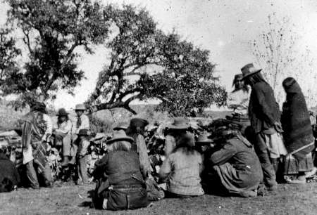 A group of Indigenous people, some sitting and some standing, gathered outdoors under large trees, dressed in traditional and Western clothing.