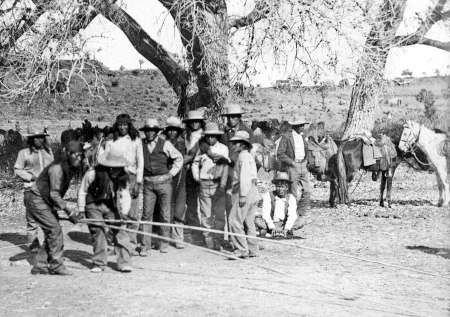 A group of people stand outdoors near trees and horses, with two individuals in the foreground pulling a rope. The setting appears rural and dry.