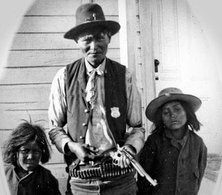A man in a vest and hat holding a revolver stands between two children in front of a wooden building.