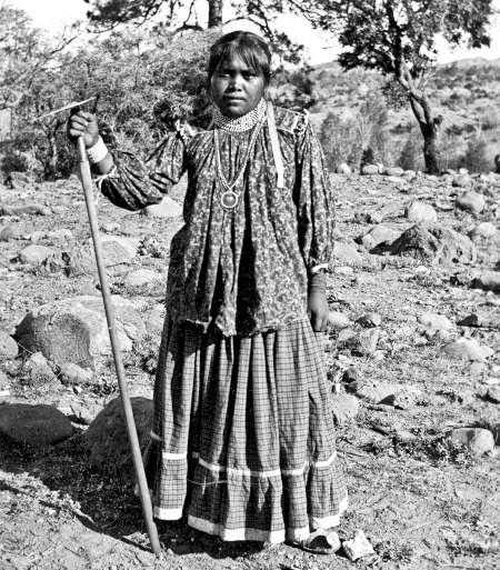 A woman in traditional dress stands outdoors on rocky terrain, holding a long stick, with trees and shrubs in the background.