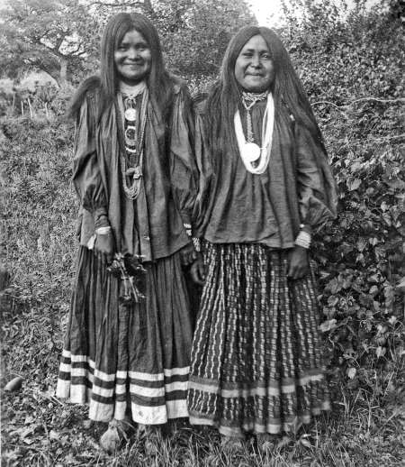 Two women in traditional clothing stand outdoors, wearing long skirts, blouses, necklaces, and long hair, with trees and foliage in the background.