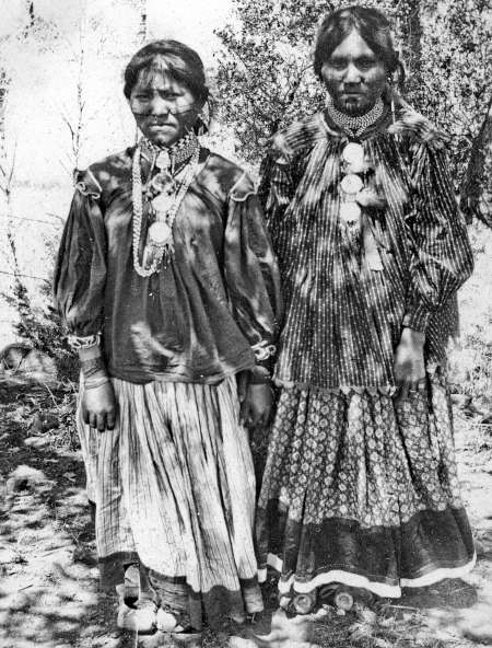Two Indigenous women stand side by side outdoors, wearing patterned dresses, necklaces, and traditional jewelry, facing the camera in a black and white photograph.
