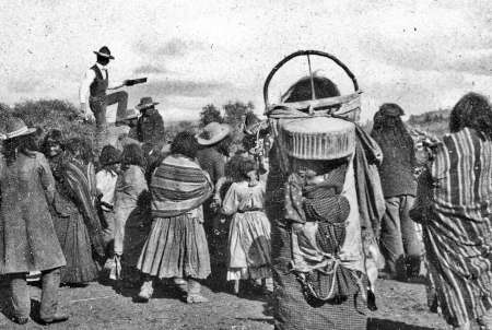A group of Indigenous people gather outdoors; a man stands elevated speaking, while a woman with a cradleboard stands in the foreground.