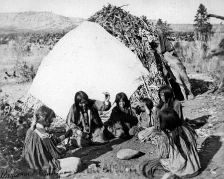 A group of Indigenous women and children sit on the ground outside a brush shelter, grinding food with stones in a natural, outdoor setting.