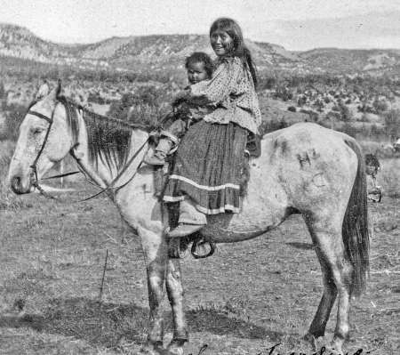 A woman and a child sit together on a horse in a grassy, open landscape with hills in the background.