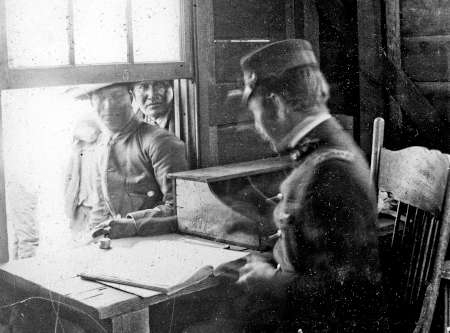 A uniformed officer sits at a desk with a ledger, while two men in hats stand at a window outside, looking in.