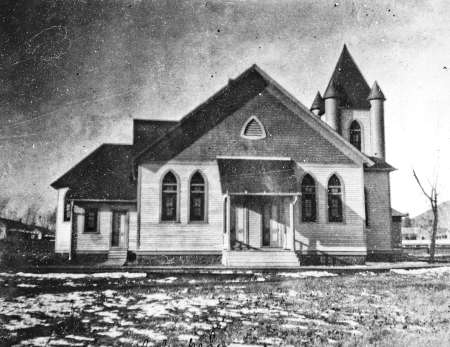 Black and white photo of a wooden church with pointed steeple, arched windows, and snow patches on the ground in front.