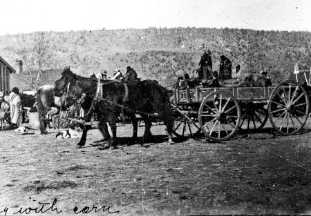 A horse-drawn wagon loaded with goods stands on a dirt area with several people nearby; hills are visible in the background.