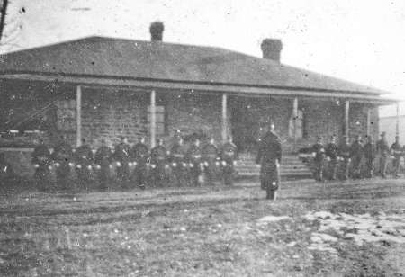 A group of soldiers is seated in front of a stone building with a porch, while one individual stands facing them on a grassy area.