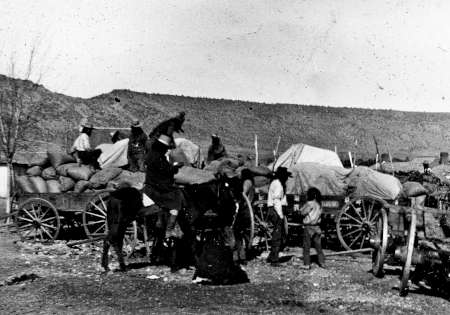 Black-and-white photo of people loading or unloading supplies from covered wagons, with a person on horseback in the foreground and hills in the background.