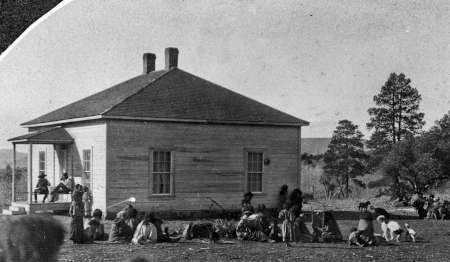 Black and white photo of a group of people gathered outside a small, wood-framed building with a porch, surrounded by open land and trees.
