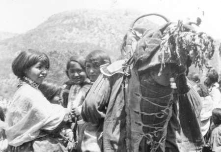 A group of children stand outdoors near a person carrying a large bundle on their back, with a mountain visible in the background.