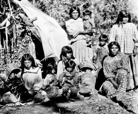 A group of Native American women and children pose outdoors beside a makeshift shelter made of branches and cloth.