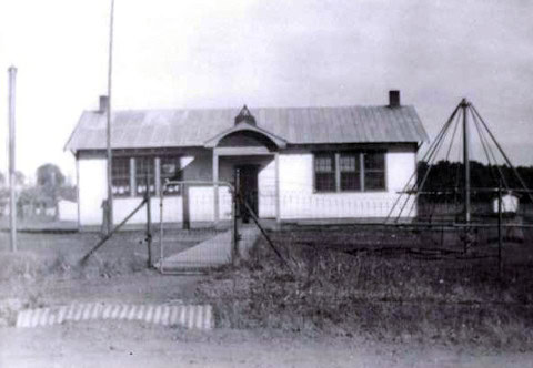 Black and white photo of a small, old-fashioned school building with a pitched roof, large windows, and a playground swing set in the foreground.