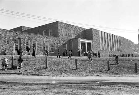 Black and white photo of people walking outside a large, rectangular stone building with tall windows and a main entrance.