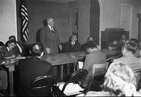 A man in a suit stands and speaks at the head of a table while several people sit and listen in a meeting room with an American flag in the background.