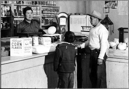 A man and a child stand at a grocery store counter talking to a shopkeeper. Food items and produce are displayed on the counter and shelves behind.