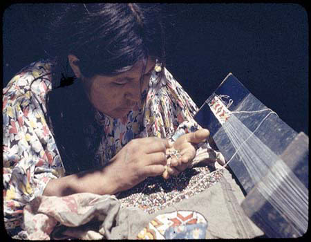 A woman in a patterned dress threads colorful beads onto a loom, focusing intently on her beadwork.